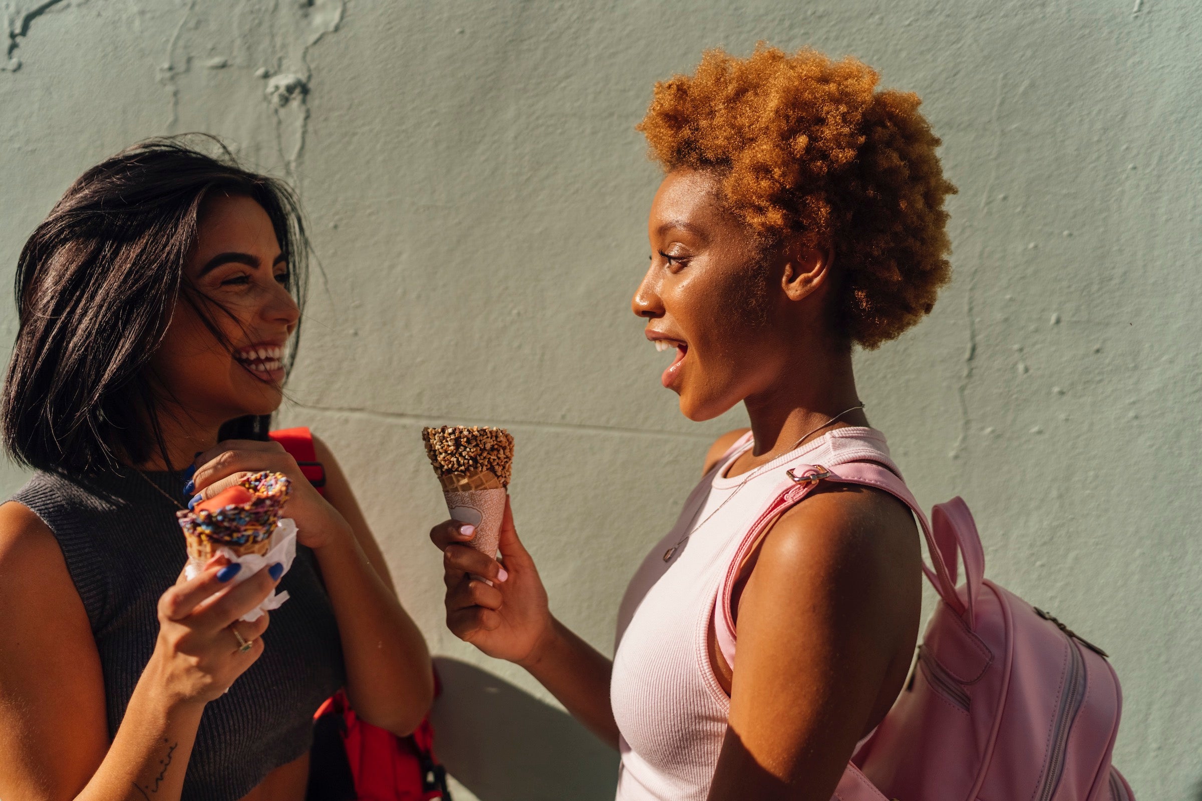Two happy female friends with ice cream cones talking at a wall
