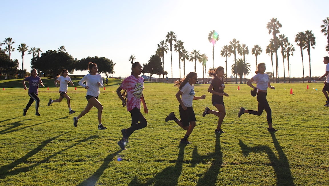 Wings of America youth runners doing strides against sunny landscape with palm trees.