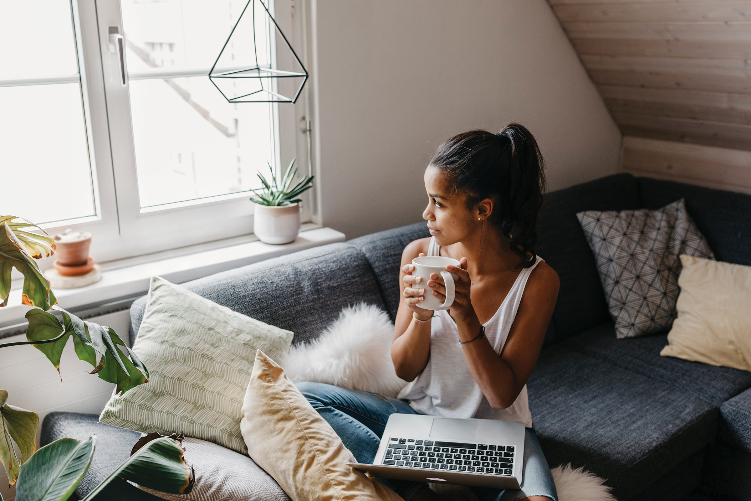 Young woman with laptop and cup of coffee sitting on the couch at home looking out of window