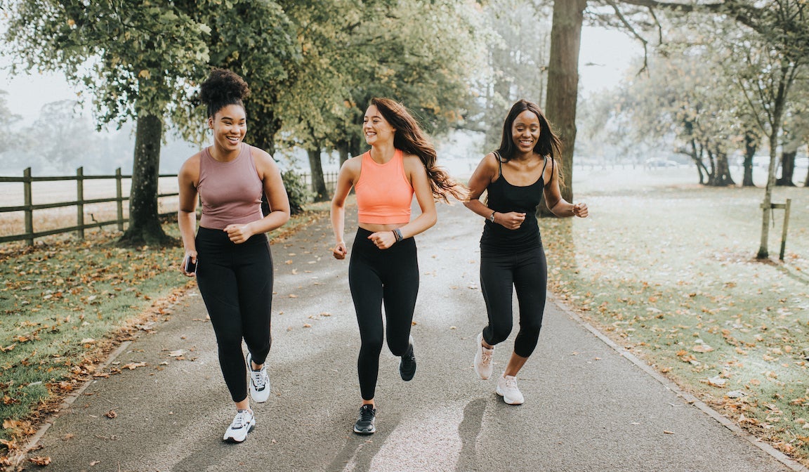 Strong, fit woman joggers, running through a sunny park. They chat and laugh as they exercise with each other. Their hair blows behind them as they look in front of them. They run towards the camera.