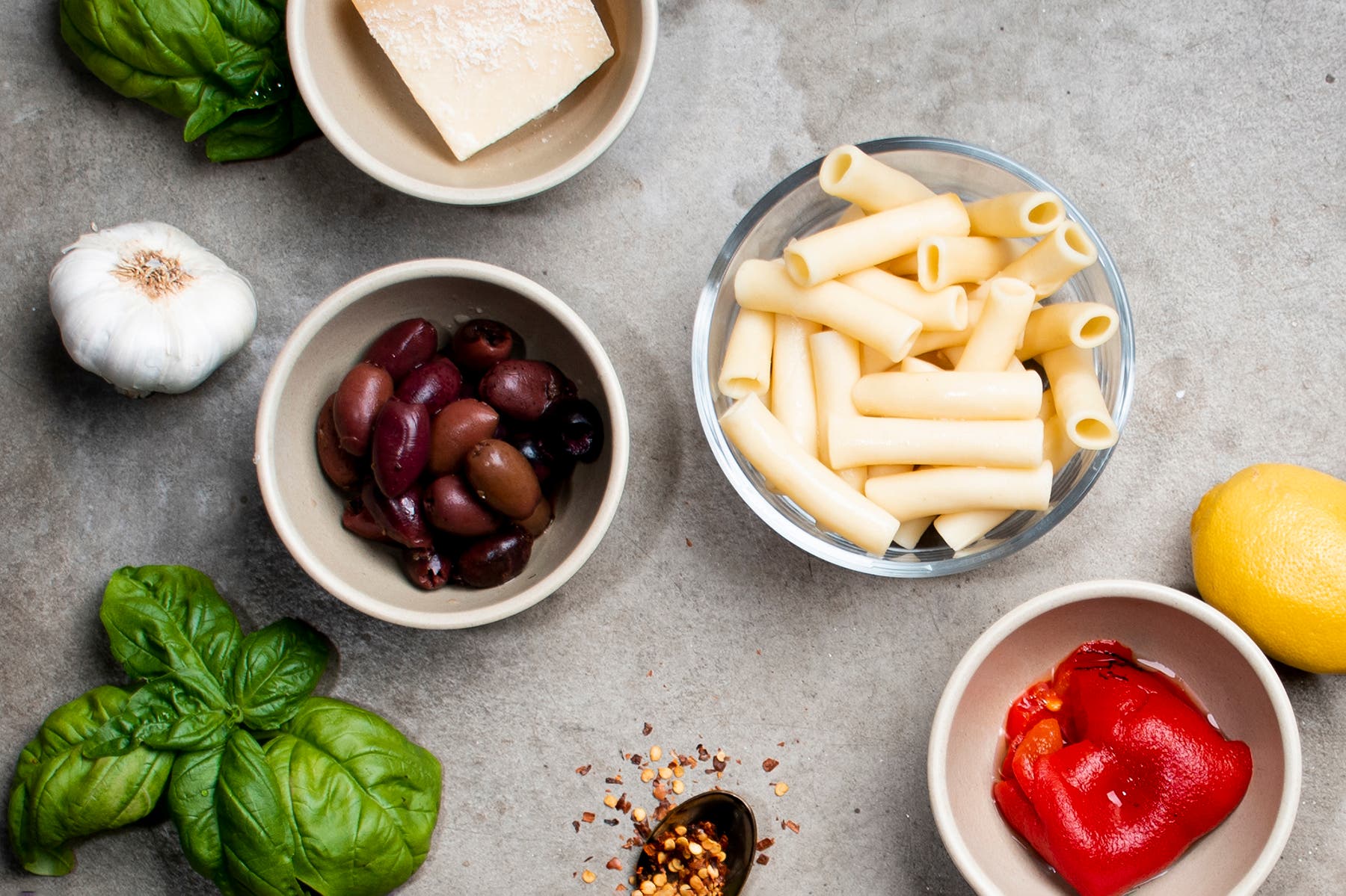 Several of the ingredients for a healthy baked ziti pasta layed out, including feta, pasta, olives, garlic, and roasted red pepper